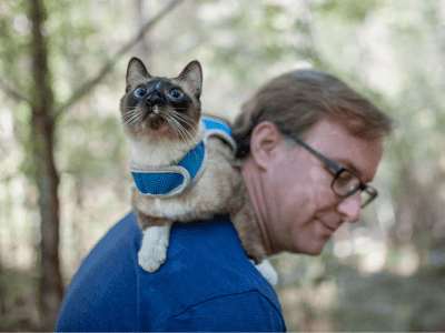 man with siamese cat on his shoulders, outside in the woods